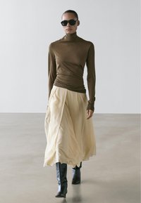 Bruine coltrui met lange mouwen, gecombineerd met een vloeiende, crèmekleurige rok, aangevuld met zwarte kniehoge laarzen en donkere zonnebril.