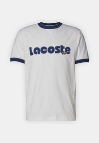 Camiseta de algodón blanca con cuello y mangas en azul marino. Presenta un gran logo azul de "Lacoste" y un emblema de cocodrilo verde en el pecho.