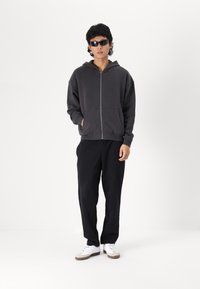 Pelēks kapuces jaka ar rāvējslēdzēju, priekšējām kabatām, brīvā piegriezuma; saskaņota ar melnām biksēm un baltām sporta apaviem. Modelis valkā tumšas saulesbrilles.