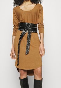 Robe longue marron à manches longues avec un col arrondi, associée à une ceinture en cuir noir ornée d'œillets en argent et de longues queues.