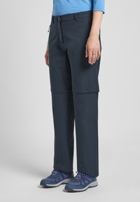 Waterdichte marineblauwe softshellbroek met ritszakken, verstelbare manchetten en een recht pijpdesign, gecombineerd met grijze sportschoenen.