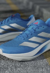 Chaussures de course Adidas bleues avec des bandes blanches et des semelles rembourrées, placées sur une surface asphaltée texturée.