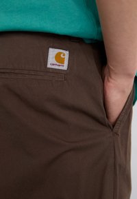 Pantaloni din bumbac maro cu detalii de buzunare, având un patch cu logo-ul Carhartt pe un fundal alb și accente galbene. Textură netedă.