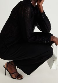 Svart spetsblus med långa, genomskinliga ärmar, tillsammans med svarta vida byxor och svarta sandaletter med klack. Håller en liten vit väska.