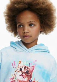 Lichtblauwe hoodie van zachte stof met een prominente afbeelding van een kattenkop, vergezeld van kleurrijke accenten en speelse tekstelementen.