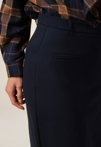 Marineblauer Bleistiftrock mit glatter Textur, ausgestattet mit einer Seitentasche und einer figurbetonten Silhouette, kombiniert mit einem karierten Hemd in Braun und Blau.