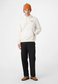 Cremefarbener Hoodie mit Kordelzug, Kängurutasche und rotem Schriftzug; kombiniert mit schwarzen Hosen und braunen Sneakern; dazu eine orange gerippte Mütze.