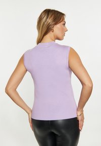 Top smanicato in maglia lavanda con scollatura alta, design aderente e texture liscia. Indossato con pantaloni neri aderenti.