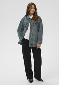 Jeansjacke mit verwaschenem blauem Finish, zwei Vordertaschen und Knopfverschluss, kombiniert mit schwarzen weiten Hosen und spitzen schwarzen Schuhen.