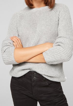 Personne portant un pull gris clair en maille avec les manches retroussées et un pantalon noir, les bras croisés à la taille.