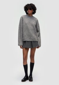 Grauer, oversized Wollpullover mit langen Ärmeln, kombiniert mit einem grauen Faltenrock. Schwarze kniehohe Socken und schwarze Slipper runden das Outfit ab.
