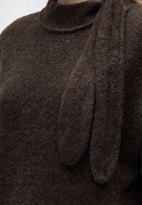 Pull marron à texture duveteuse avec un col montant et une paire de longues extensions en tissu doux, en forme d’oreilles, suspendues d’un côté.