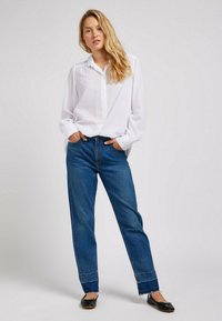 Camicia bianca con bottoni e maniche lunghe, abbinata a jeans blu a gamba dritta con orli sfrangiati e ballerine nere.