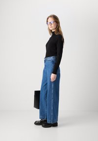 Svart långärmad topp, högmidjade vida blå jeans, svarta platåskor och glasögon med ljuslila ton; håller en svart väska.