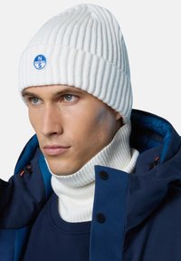 Beanie a coste bianco con un logo circolare blu. Il modello indossa un dolcevita bianco a coste e un giaccone blu navy con bottoni.