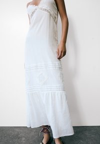 Robe blanche longue jusqu'au sol en tissu tissé texturé, avec des accents en dentelle et un ourlet en volants superposés. Elle présente des bretelles fines et une broderie géométrique.