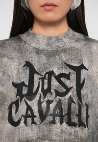 Sweatshirt cinza com um padrão texturizado e desgastado, apresentando letras pretas em negrito que dizem "JUST CAVALLI." Gola alta e mangas longas.