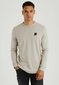 Beige Langarmshirt mit rundem Halsausschnitt, gerippter Textur und einem kleinen schwarzen Logo-Patch auf der Brust, kombiniert mit dunklen Jeans.