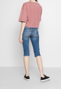 Camiseta corta de manga corta en rosa empolvado combinada con pantalones cortos de mezclilla hasta la rodilla. Los pantalones cortos presentan un lavado azul desgastado y dobladillos rematados. Zapatillas negras.