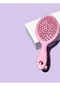 Brosse à cheveux en plastique rose de forme ronde, dotée d'une grille de poils flexibles et d'un manche ergonomique prononcé. Placée sur une boîte blanche.