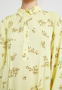Blusa giallo pallido con piccoli motivi floreali marroni, apertura frontale con quattro bottoni e pieghe sottili lungo la zona del petto.