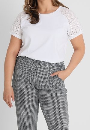 Femme portant une chemise blanche à manches courtes en dentelle et un pantalon gris à motifs avec une ceinture nouée, une main dans la poche.