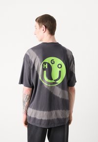Pilkas t-shirt su rašteliu, dekoruotas išlenktu neoninės žalios spalvos logotipu ir "HGO" tekstu ant nugaros. Trumpomis rankovėmis, laisvas pritaikymas ir minkšta audinio tekstūra.