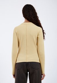 Woman with long curly hair wearing a textured beige long-sleeve top and dark high-waisted jeans, shown from the back against a white background.