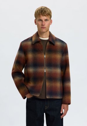 Jeune homme aux cheveux blonds courts portant une veste à carreaux marron, orange et noire zippée et un jean foncé sur un fond uni.
