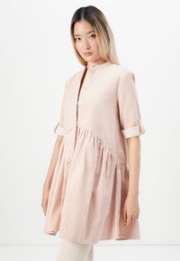 Noor noor, seljas heledat roosat, nööpidega vabalt langevat kleiti, mille varrukad on üles keeratud, lihtsa valge tausta ees.