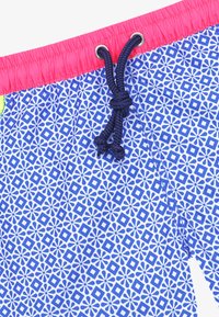 Maillots de bain bleus à motifs avec une taille rose. Présente des motifs floraux géométriques et un cordon de serrage bleu marine pour l'ajustement.