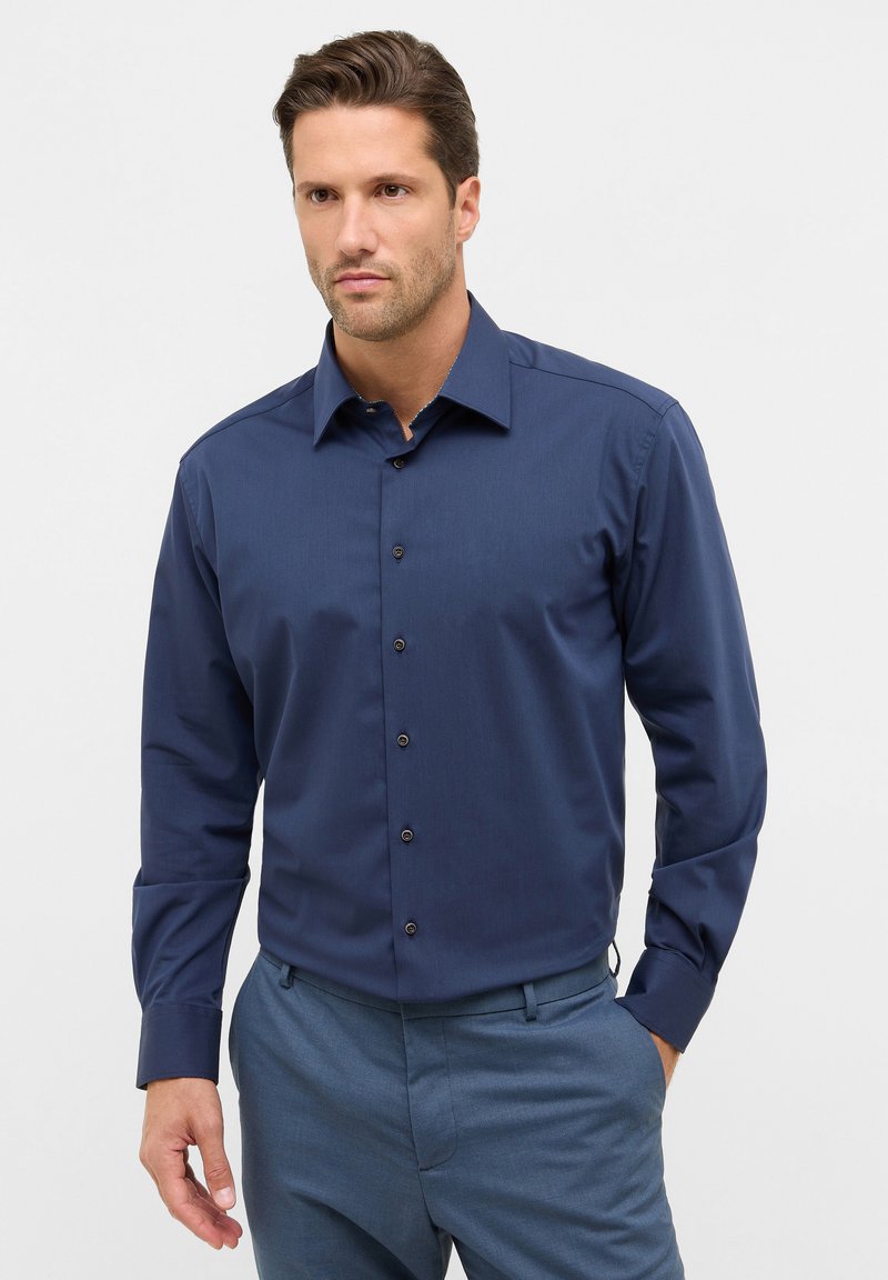 Camicia navy blue con bottoni e maniche lunghe, dalla trama sottile e bottoni neri. Indossata con pantaloni azzurri. Aspetto pulito e sartoriale.