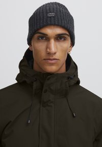 Beanie grigio a coste con una toppa con il marchio; giacca impermeabile verde scuro con colletto alto, lacci e bottoni a scatto; texture liscia.