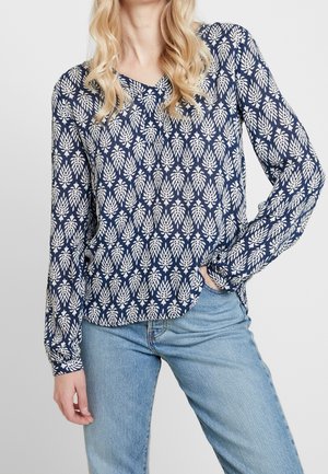 Femme blonde portant un chemisier bleu marine à manches longues avec des motifs de feuilles blanches et un jean bleu clair, une main dans la poche.