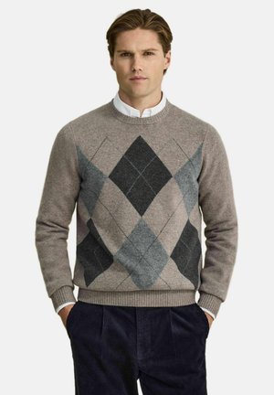 Mand iført en grå og sort argyle-mønstret sweater over en hvid skjorte med krave, med marineblå fløjlsbukser, hænder i lommerne.