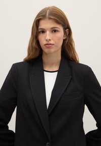 Blazer nero con un bottone singolo, rever a intaglio e una tasca sul petto. Indossato sopra una maglia bianca con un dettaglio nero. Texture liscia.