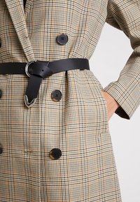 Dvojřadový plaid kabát v béžové a hnědé barvě s černým pásem. Obsahuje velké knoflíky a volný střih, vyrobený z texturované látky.