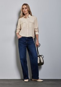 Camicia button-up beige chiaro con due tasche sul petto, abbinata a jeans a gamba larga blu scuro. Il modello tiene una borsa piccola, indossando ballerine beige.