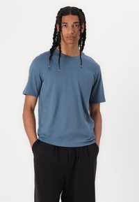 Blue short-sleeve T-shirt in a smooth, stretchy fabric with a relaxed fit. Worn with black trousers, featuring a crew neckline.