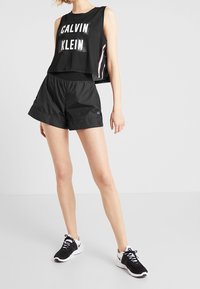 Nainen, joka pitää yllä mustaa Calvin Klein -hihattomaa paitaa, mustia urheilushortseja ja musta-valkoisia lenkkareita, seisoo yksinkertaisella taustalla.