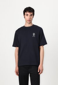 T-shirt en coton bleu marine avec un col rond, des manches courtes et une petite impression graphique sur la poitrine gauche. Coupe décontractée et texture lisse.