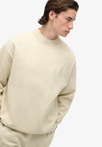 Giovane uomo con capelli scuri che indossa un felpa e pantaloni beige coordinati, che guarda di lato con le mani in tasca su uno sfondo bianco.