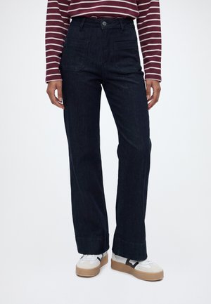 Person iført mørke vide jeans, hvide sneakers med beige såler og en langærmet bordeaux og hvide stribet trøje.
