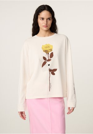 Vrouw met lang donker haar, dragen een crèmekleurig shirt met lange mouwen met een gele roos en een lichtroze rok tegen een effen achtergrond.
