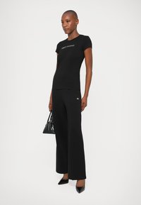 Camiseta negra ajustada con el logo de "Armani Exchange", combinada con pantalones de pierna ancha negros de talle alto. Accesorios incluyen un pequeño bolso negro.