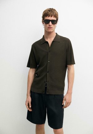 Junger Mann, der ein dunkelgrünes kurzärmeliges Hemd mit Knöpfen, schwarze Shorts und schwarze Sonnenbrillen trägt, steht vor einem schlichten weißen Hintergrund.