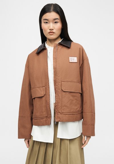 Γυναίκα που φοράει καφέ μπουφάν με μεγάλες μπροστινές τσέπες και μαύρο γιακά πάνω από λευκό πουκάμισο και μπεζ πλισέ φούστα, με φόντο λευκό.