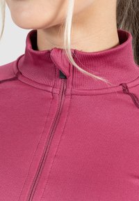 Upravený, vroubkovaný, dlouhý rukáv top v sytě růžové barvě. Obsahuje vysoký límec a centrální zip s černým detailem. Viditelná struktura tkaniny.