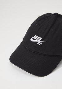 Czarna czapka bejsbolowa z materiału z wyszywanym białym logo Nike i napisem "SB" z przodu. Gładka tekstura, zakrzywiony daszek i sześciopanelowy design.