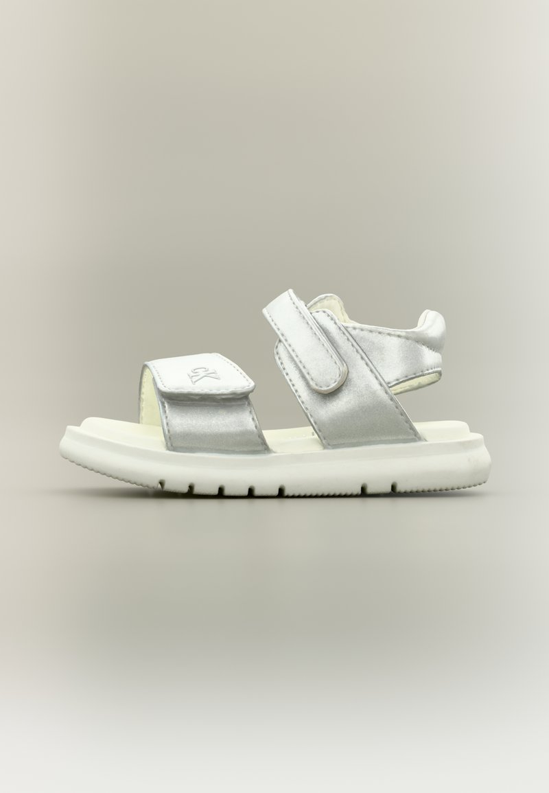 Ασημένιο και λευκό παιδικό σανδάλι με δύο ρυθμιζόμενα λουράκια με βέλκρο και χοντρή, αυλακωτή σόλα σε ουδέτερο φόντο.